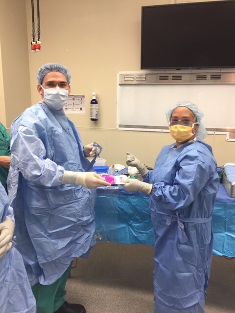 Two doctors in blue gowns and masks stand next to a table.